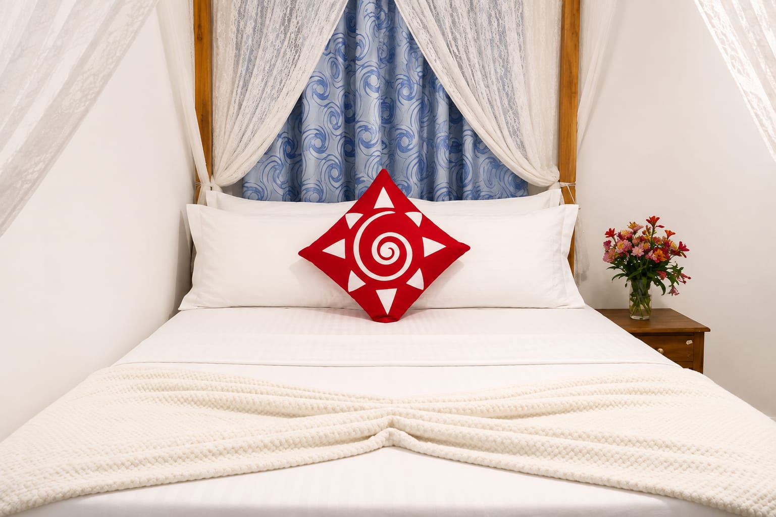 Deluxe Double Room: a four-poster bed with white linen, a red hand-printed cushion and fresh flowers on the bedside table.